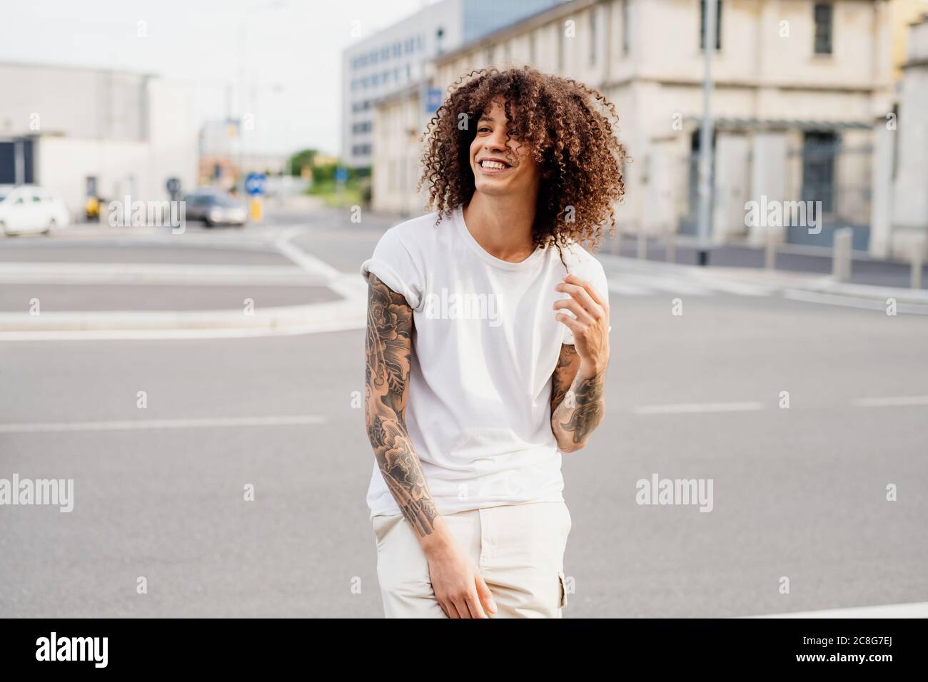Smiling man with tattooed arms and long brown curly hair standing on a street. Stock Photo