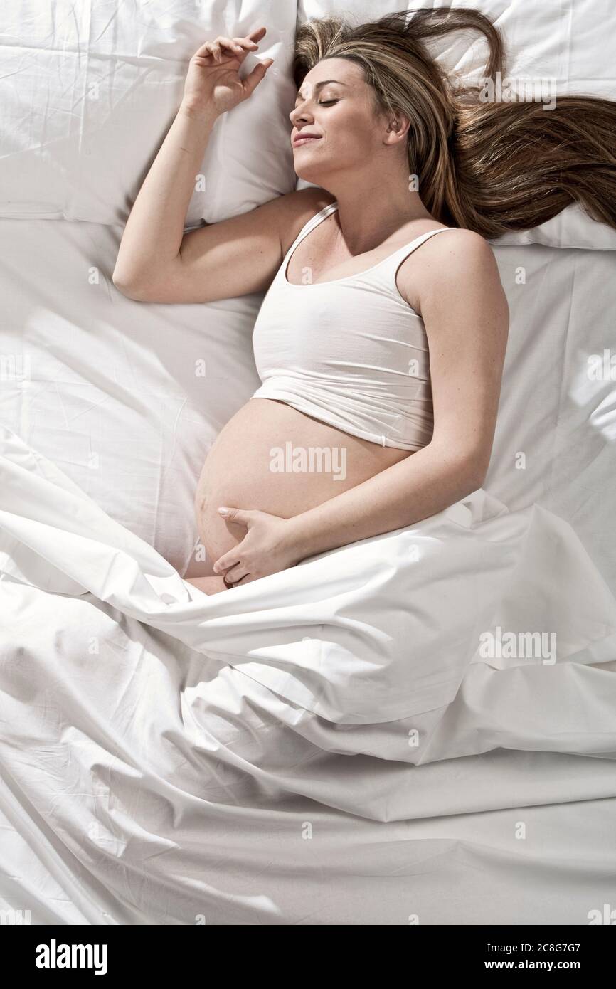 Portrait of heavily pregnant woman lying on bed, cradling stomach Stock