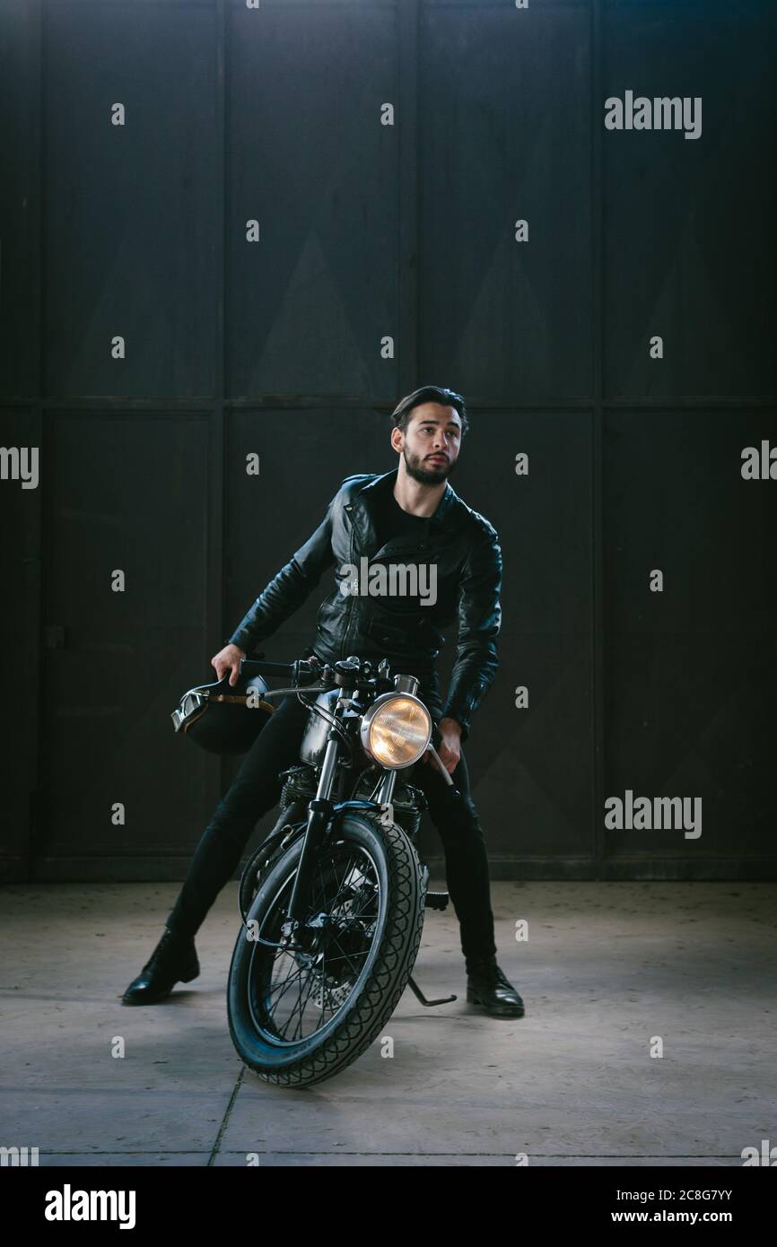 Young male motorcyclist straddling vintage motorcycle in empty warehouse Stock Photo