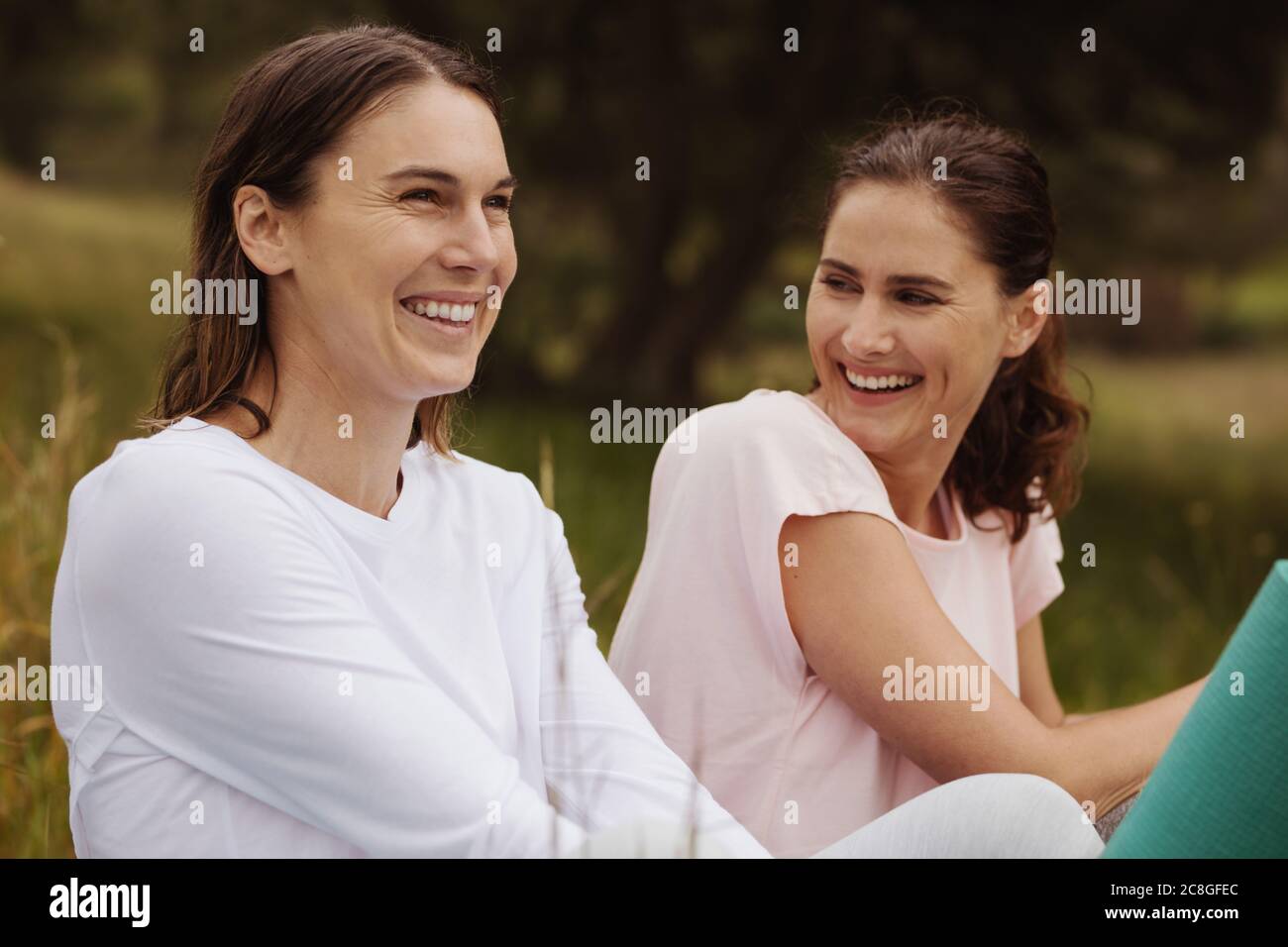 Close up of two smiling women sitting in park. Cheerful women friends sitting outdoors and looking away. Stock Photo