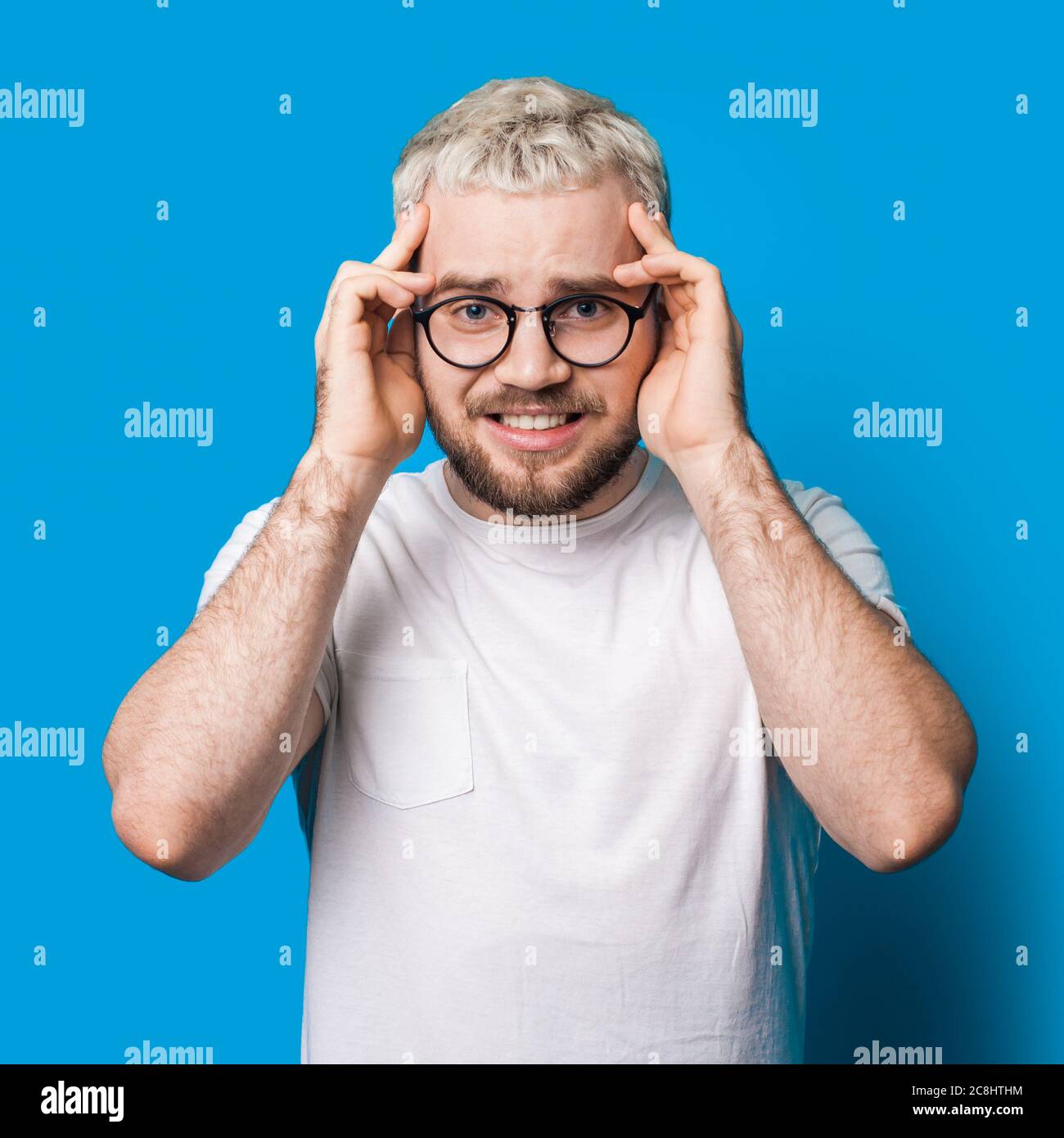Stressed bearded caucasian man with blonde hair and eyeglasses touching his  head on a blue studio wall Stock Photo - Alamy