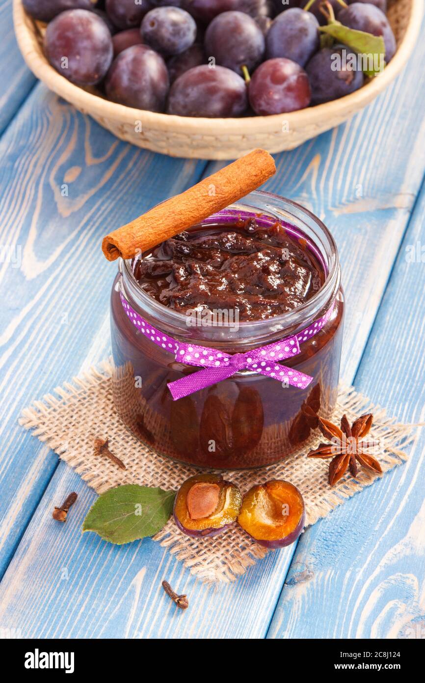 Fresh homemade plum marmalade or jam in glass jar, spices and ripe fruits in wicker basket in background, concept of healthy sweet dessert Stock Photo