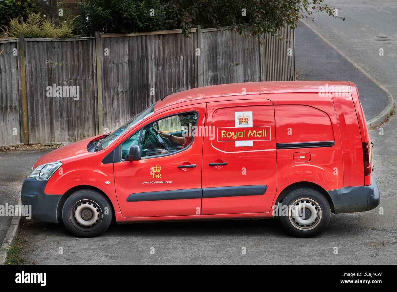 A postman sits in his bright red Royal Mail delivery van (with its royal coat of arms) after posting a package to a house. Stock Photo