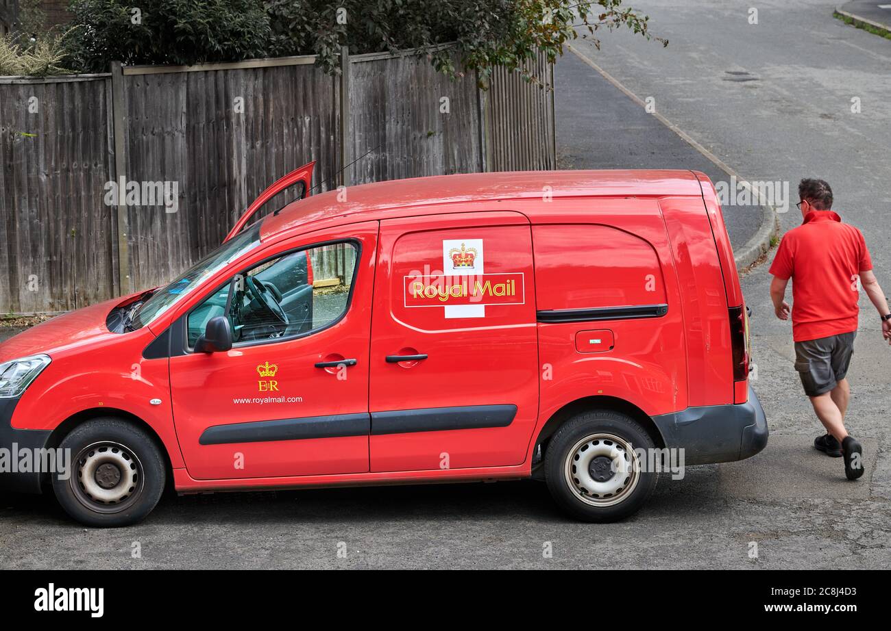 A postman returns to his bright red Royal Mail delivery van (with its royal coat of arms) after posting a package to a house. Stock Photo