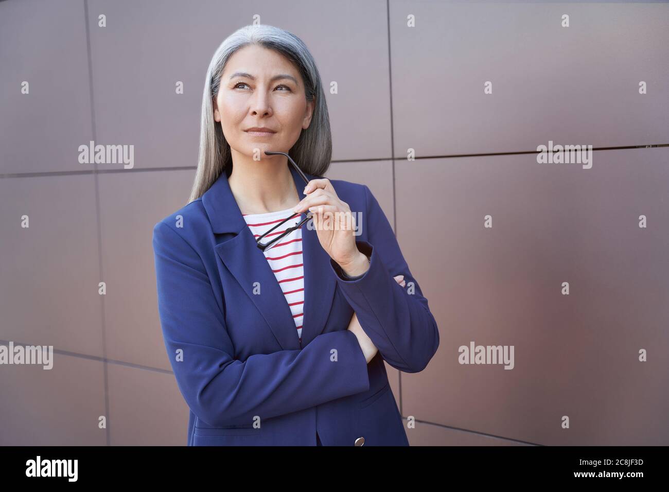 Lost in thoughts. Portrait of a serious mature business woman in classic wear holding eyeglasses, looking aside and thinking about something, standing against the wall outdoors. Business people Stock Photo