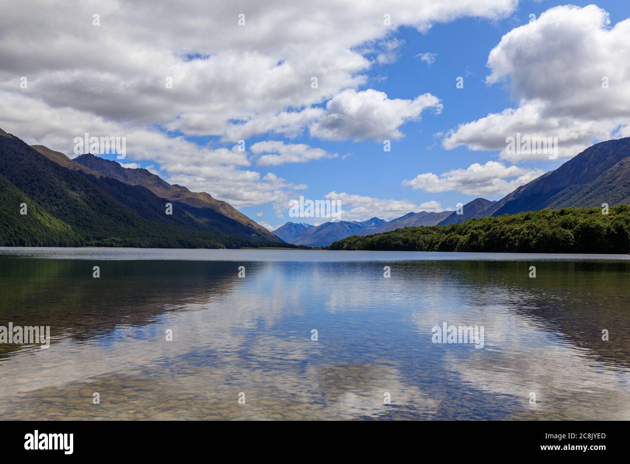 The calm waters of South Mavora Lake with forested mountains down each side and mountains in the distance. White Fluffy clouds on a blue sky. Stock Photo