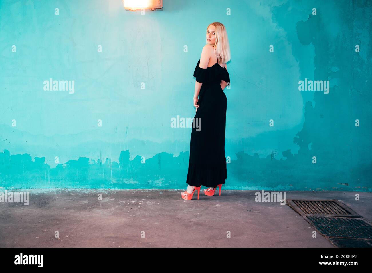 Blonde female standing in front of a light blue wall; she is wearing a long black dress with bare shoulders and high heels. Stock Photo
