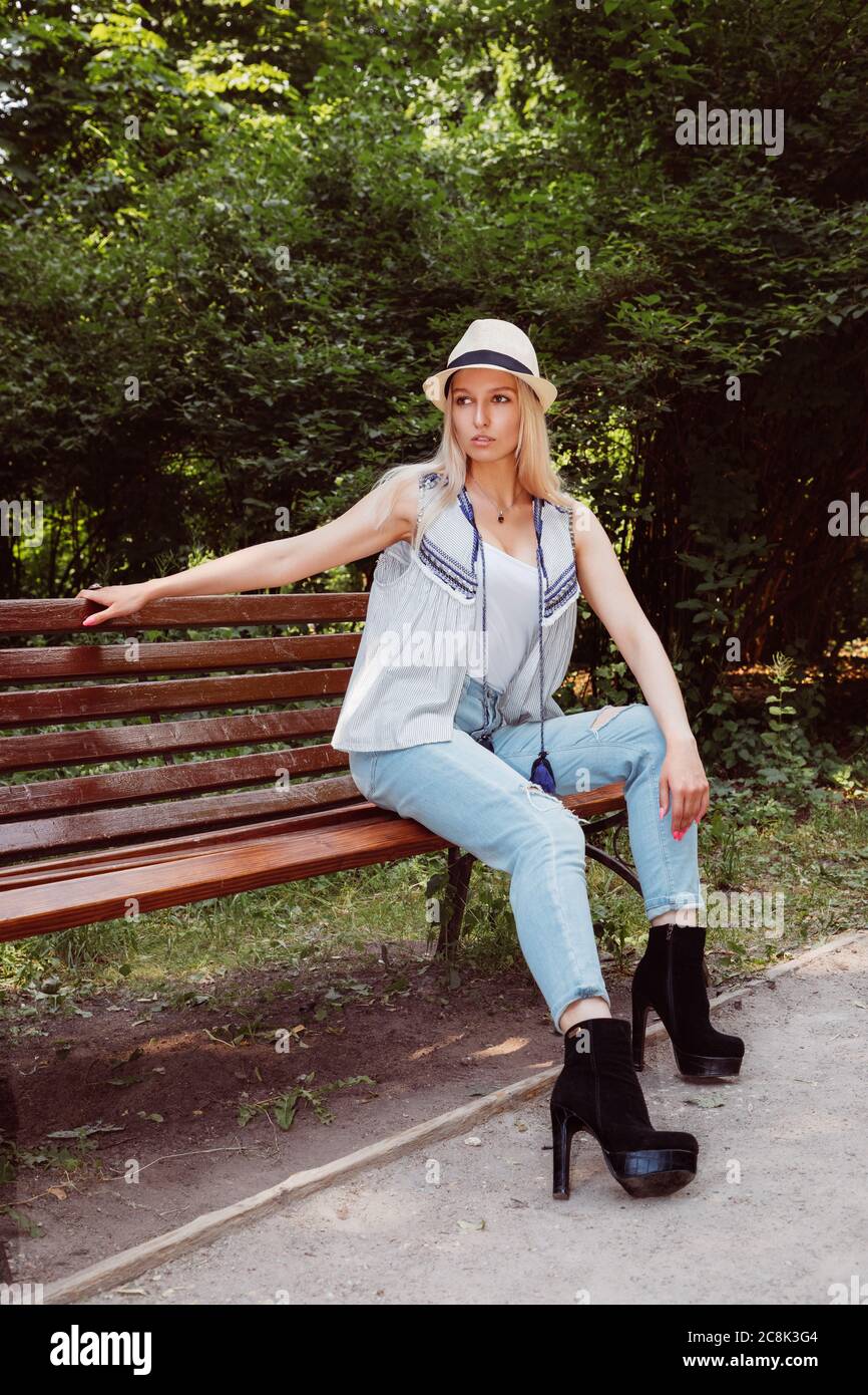 Blonde girl wearing a hat, blue jeans and boots with high heels sits on a bench in a park or forest Stock Photo