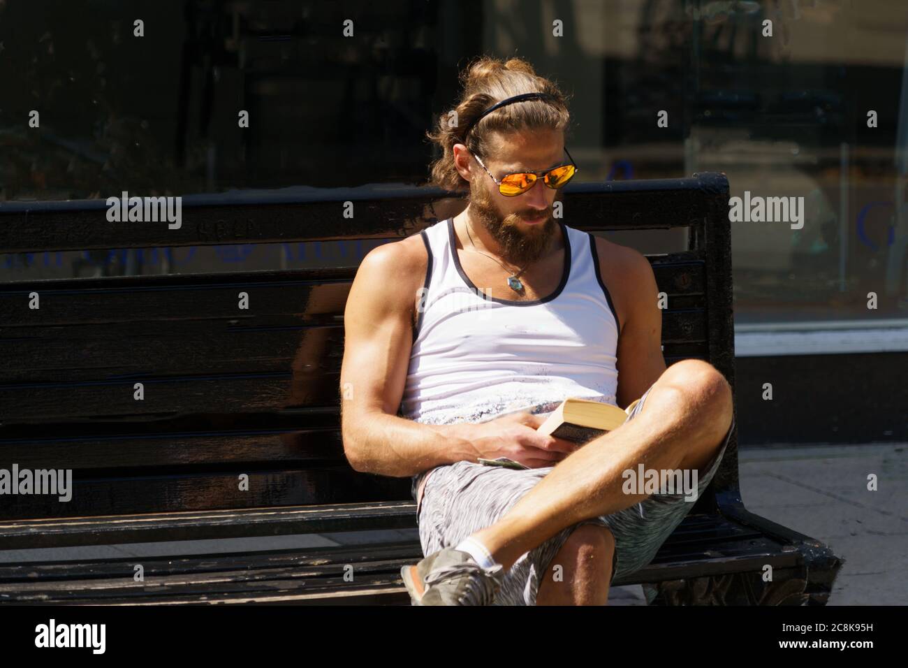 While reading a book, a man sat on a street bench wearing yellow and orange reflective sunglasses, York, North Yorkshire, England, UK. Stock Photo