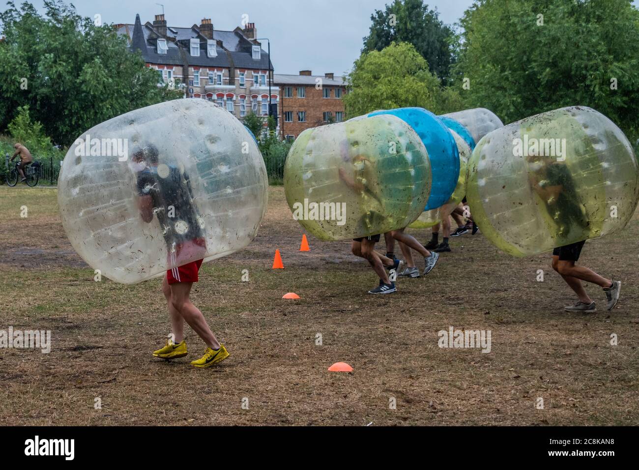 London, UK. 25th July, 2020. A new approach to social distancing partying and bubbling up, as a company called Bubble Boys oraganises an activity party for a group of men, who play british Bulldog in giant plastic balls - The first heavy rain doesn't stop outdoor parties on Clapham Common. The easing of the 'lockdown' continues for free up social activity after the Coronavirus (Covid 19) outbreak in London. Credit: Guy Bell/Alamy Live News Stock Photo