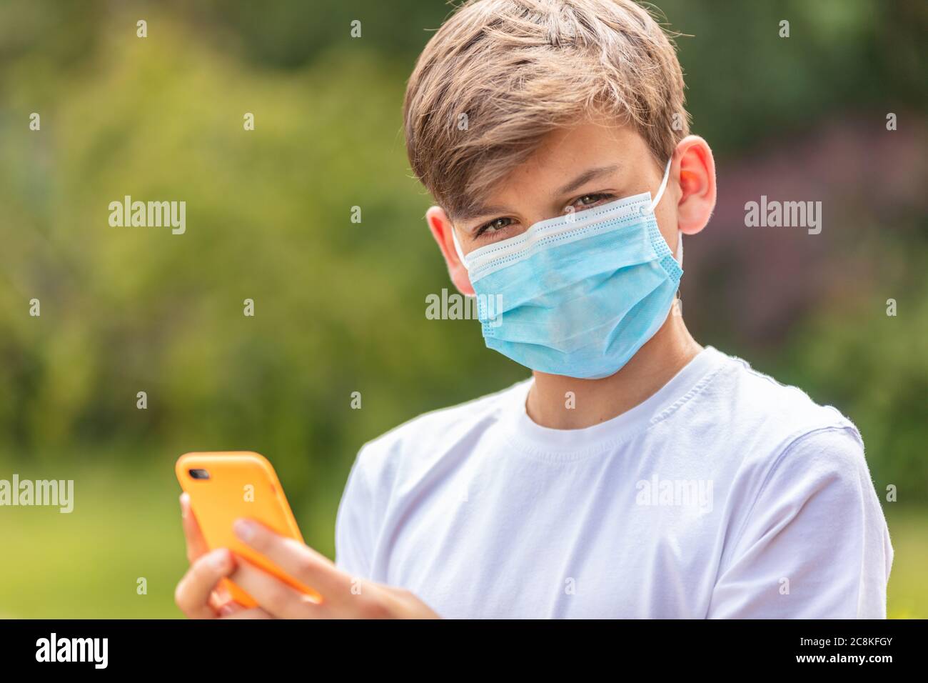 Boy teenager teen male child wearing a face mask outside during the Coronavirus COVID-19 virus pandemic while using his mobile cell phone for social m Stock Photo