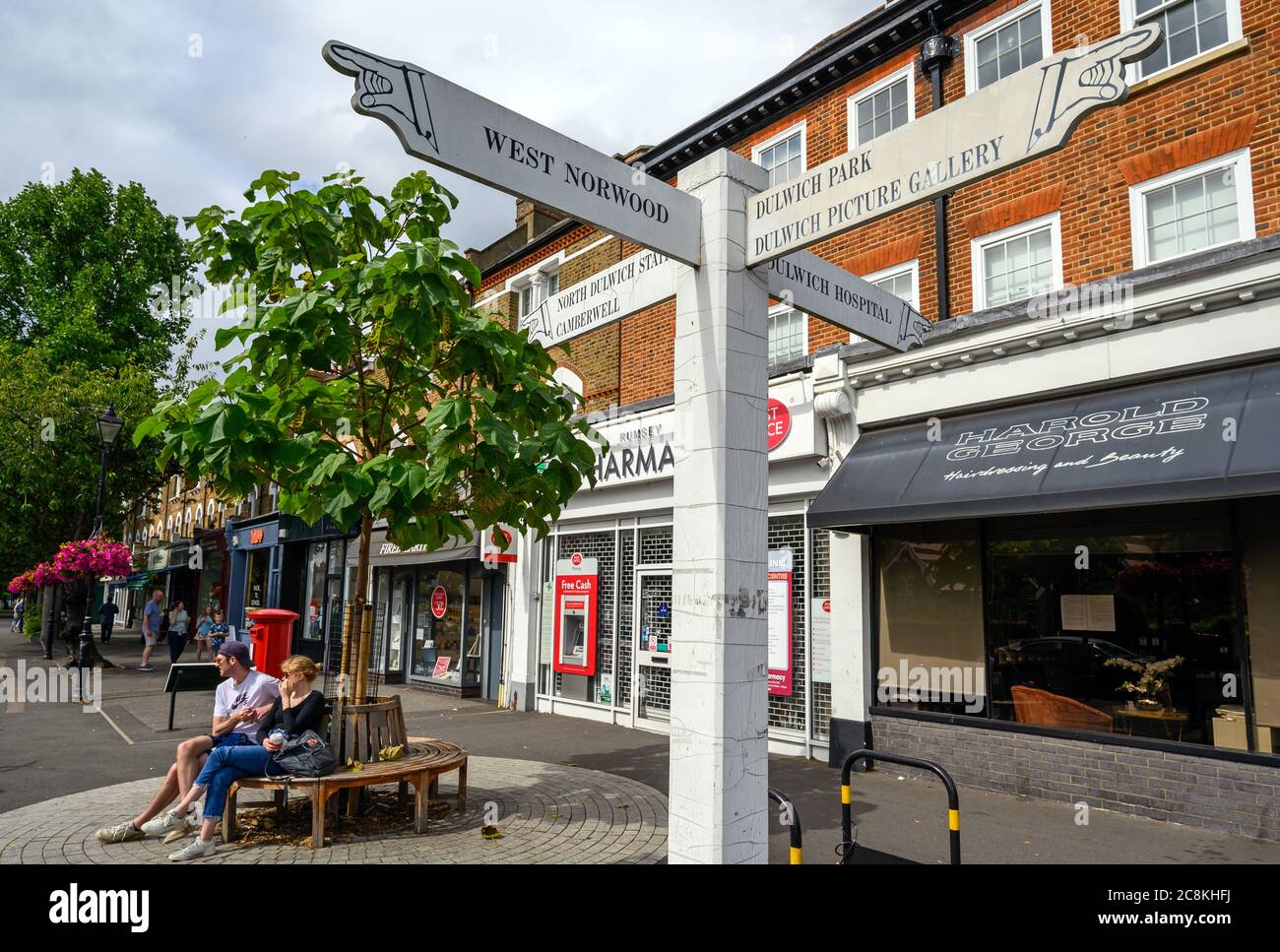 A local sign in Dulwich Village with people and shops. The sign shows directions to Dulwich landmarks and nearby places. Dulwich is in south London Stock Photo