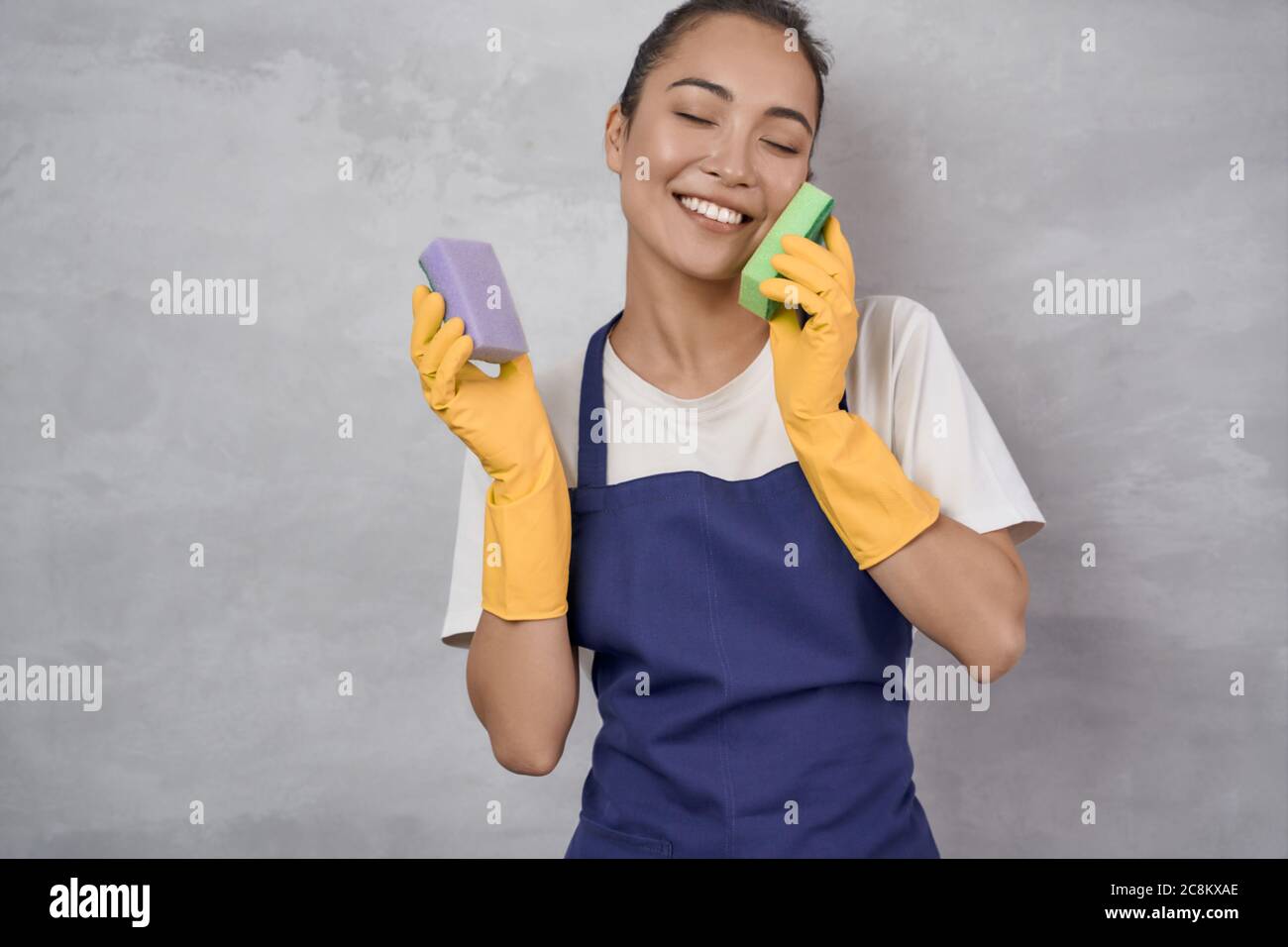 Washing dishes with fun. Happy young cleaning lady in yellow rubber gloves playing with kitchen sponges and smiling while standing against grey wall. Studio shot. Housekeeping, cleaning services Stock Photo