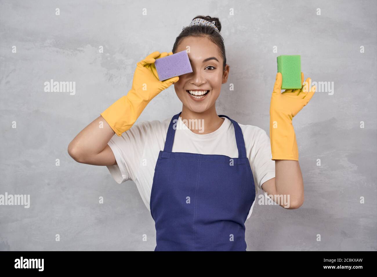 Great mood for cleaning. Funny cleaning lady in uniform and rubber gloves covering one eye with kitchen sponge for washing dishes and having fun, standing against grey wall. Housekeeping Stock Photo