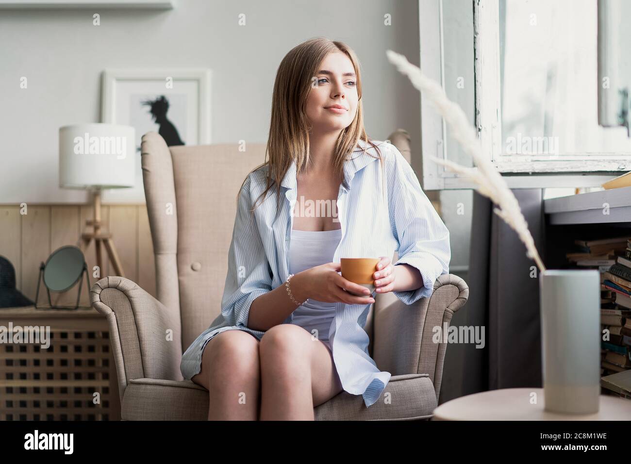 Beautiful young woman drinking coffee and looking through window while sitting at armchair at home. Stock Photo