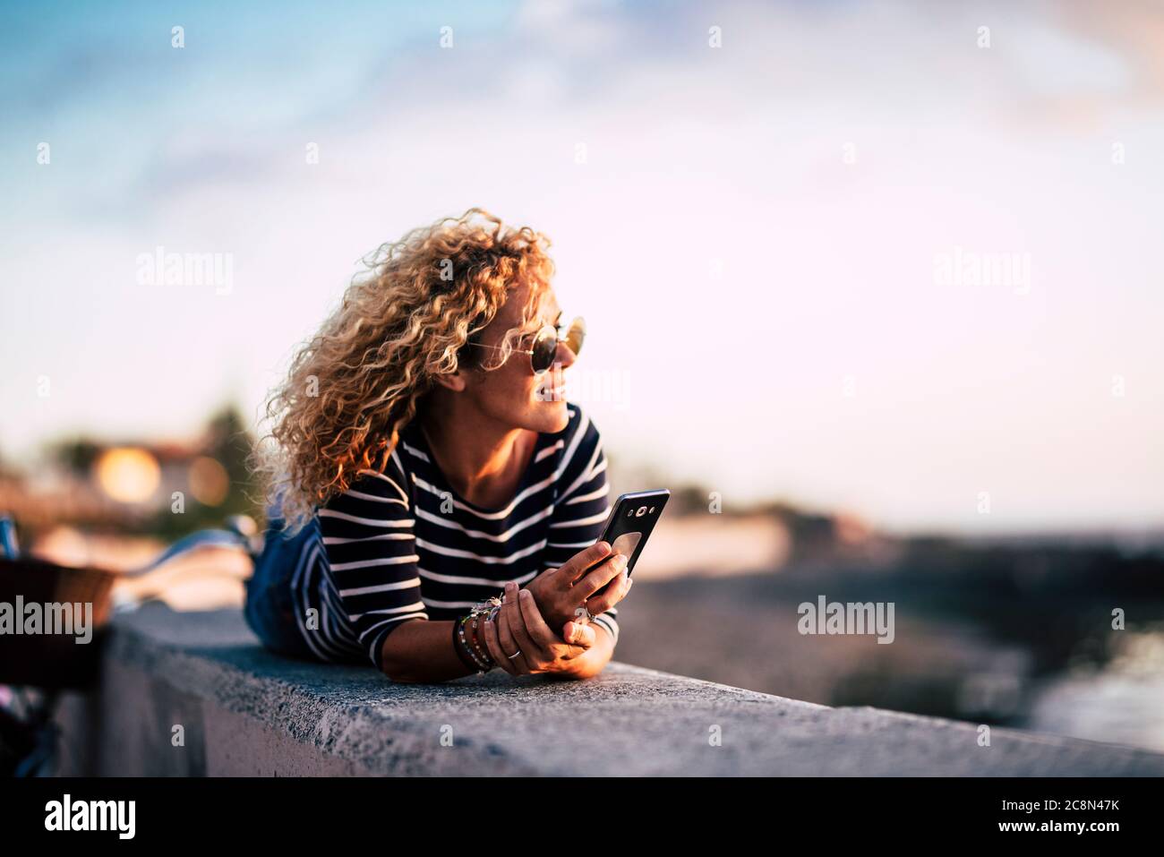 Beautiful middle age blonde curly womoan enjoy and relax in outdoor leisure activity looking at the sunset with modern phone ready to call friends - p Stock Photo