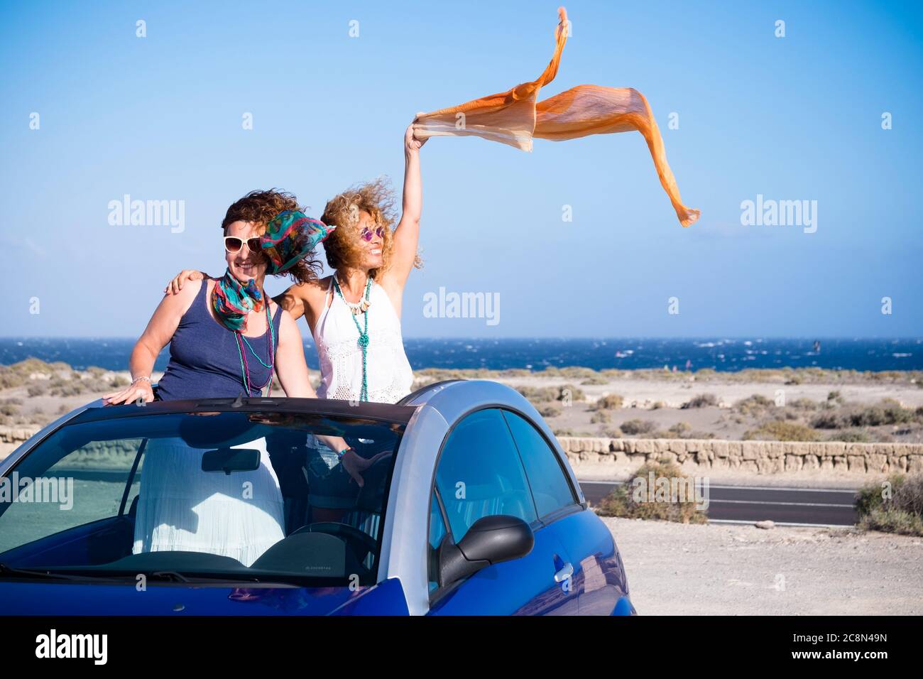 Couple of adult women friends stand up inside a convertible car enjoy the summer holiday travel vacation with vehicle - blue ocean ans sky in backgrou Stock Photo