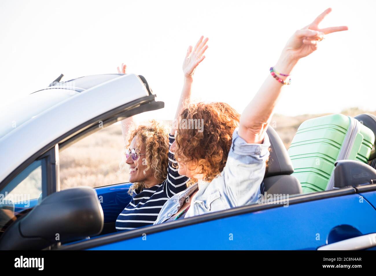 Happy travel adult friends lifestyle concept with couple of adult caucasian woman in a convertible car and luggage behind the seats - up arms and happ Stock Photo