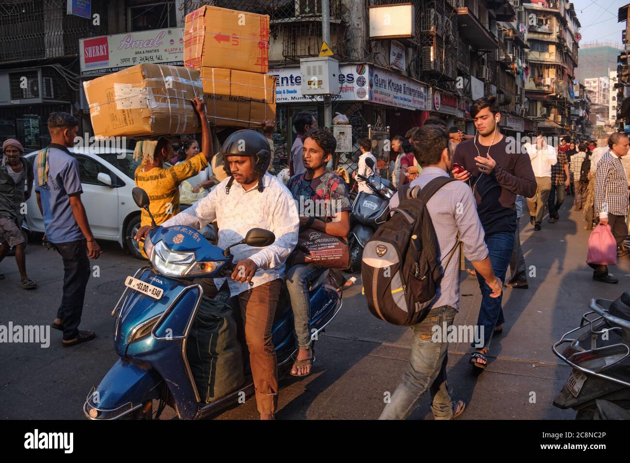 A man steers his motorcycle through busy Kalbadevi Road in Bhuleshwar, Mumbai, India, past porters with goods on their heads and numerous pedestrians Stock Photo