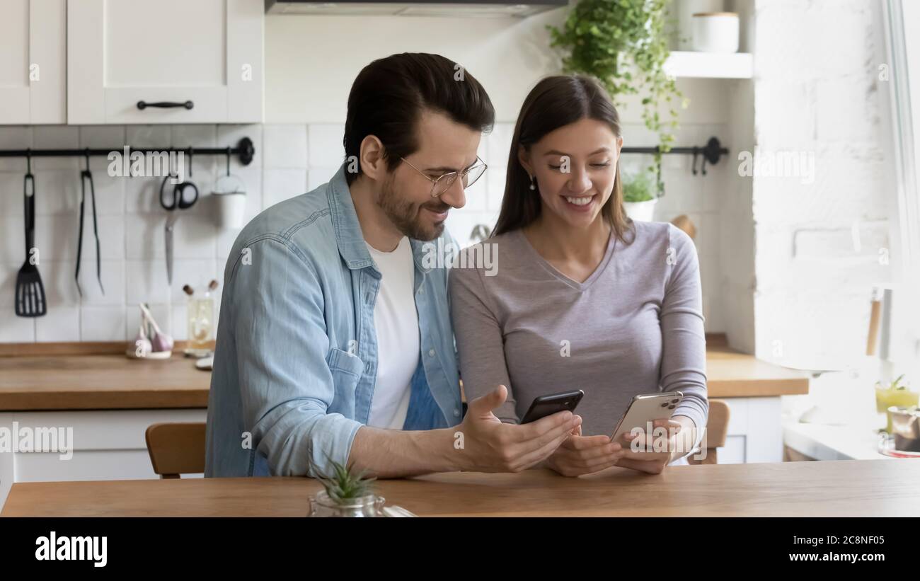 Smiling millennial couple relax on kitchen using smartphones Stock Photo