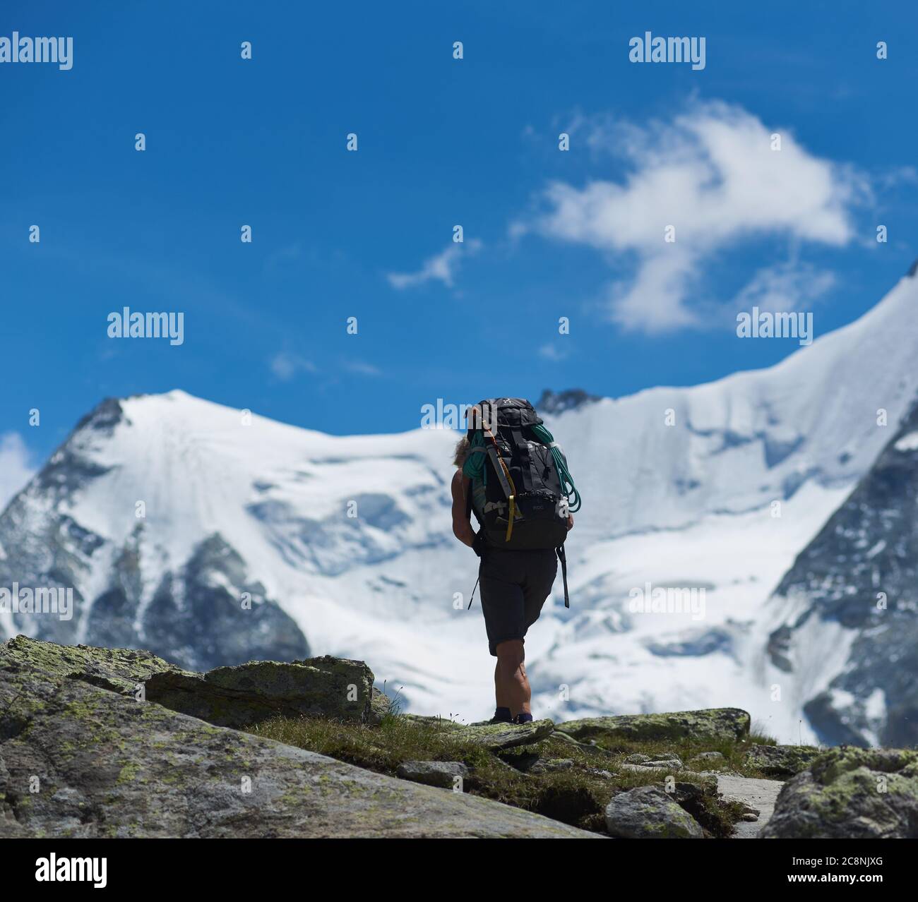 Zinal, Switzerland - July 19, 2019: Back view of traveler with backpack hiking alone in mountainous region. Backpacker walking down hillside road with snowy mountain and blue sky on background Stock Photo
