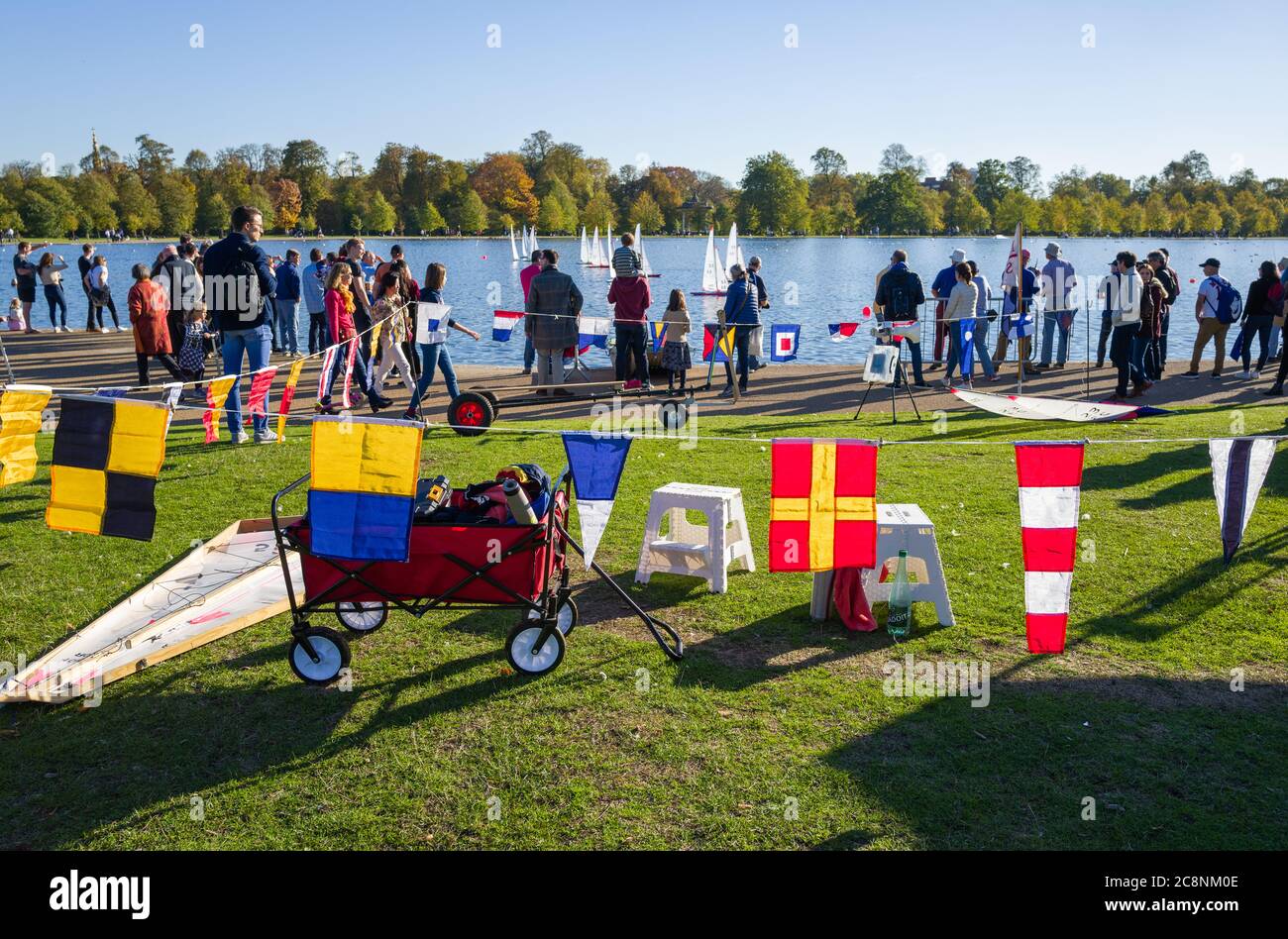 Model boats and their owners at the Serpentine in Hyde Park on a beautifully sunny and colourful autumn day. Colourful flags backlit in the foreground Stock Photo