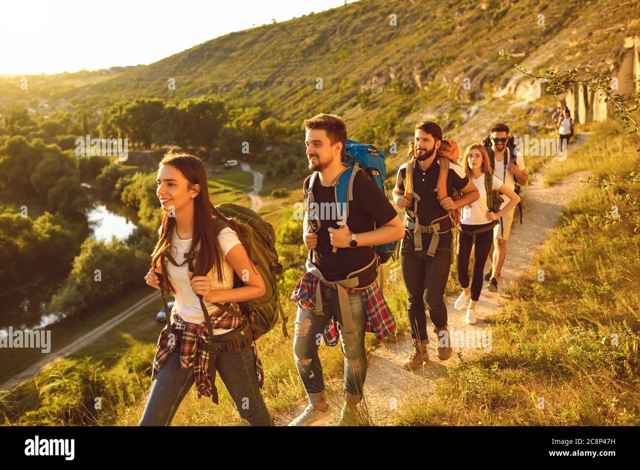 Young people with backpacks walking along narrow path in mountains. Group of cheerful tourists hiking outside in summer Stock Photo