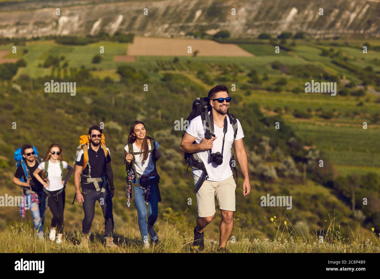 A group of friends with backpacks on a hike in nature. Young people tourists are walking on a hill in the mountains. Stock Photo