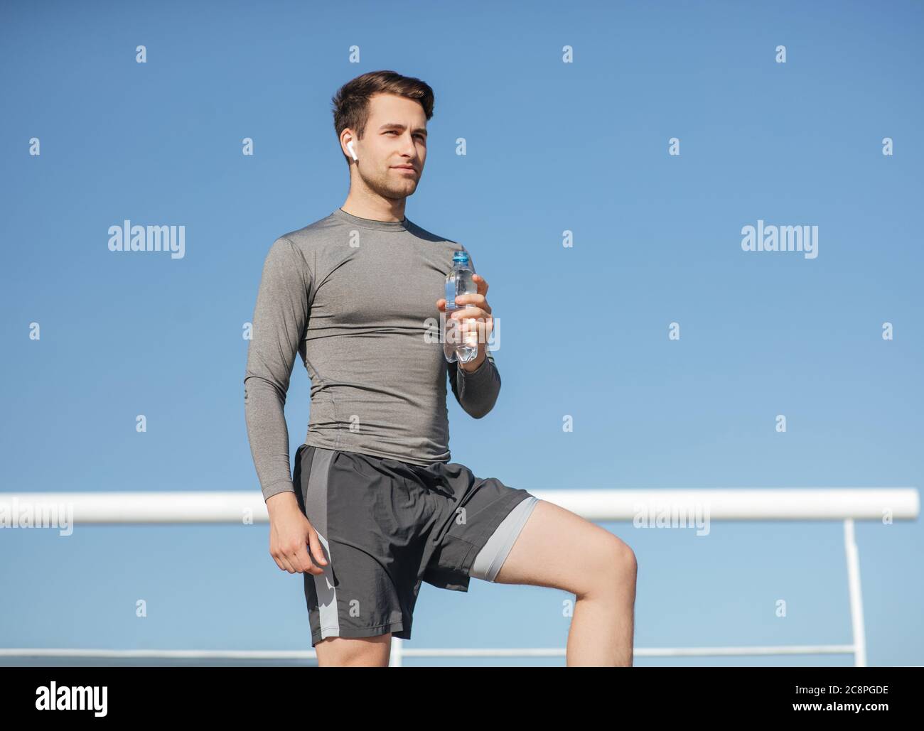Health and body care. Muscular handsome guy in sportswear, with wireless headphones holding bottle of water on blue sky background Stock Photo