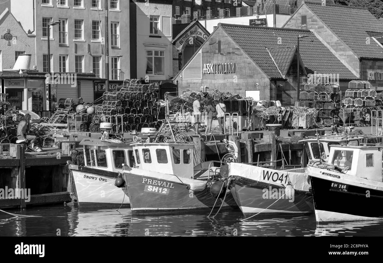 Fishing boats are moored alongside a wharf which is stacked with lobster pots. Buildings line the waterfront and tourists enjoy the surroundings. Stock Photo