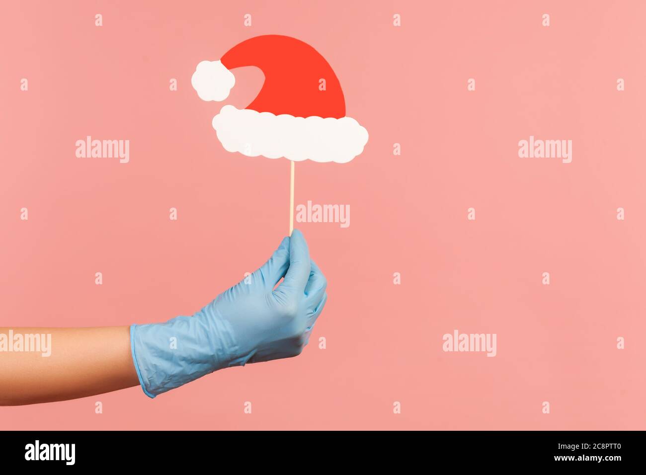 Profile side view closeup of human hand in blue surgical gloves holding and showing stick photo-booth of new year, Christmas red hat in hand. indoor, Stock Photo
