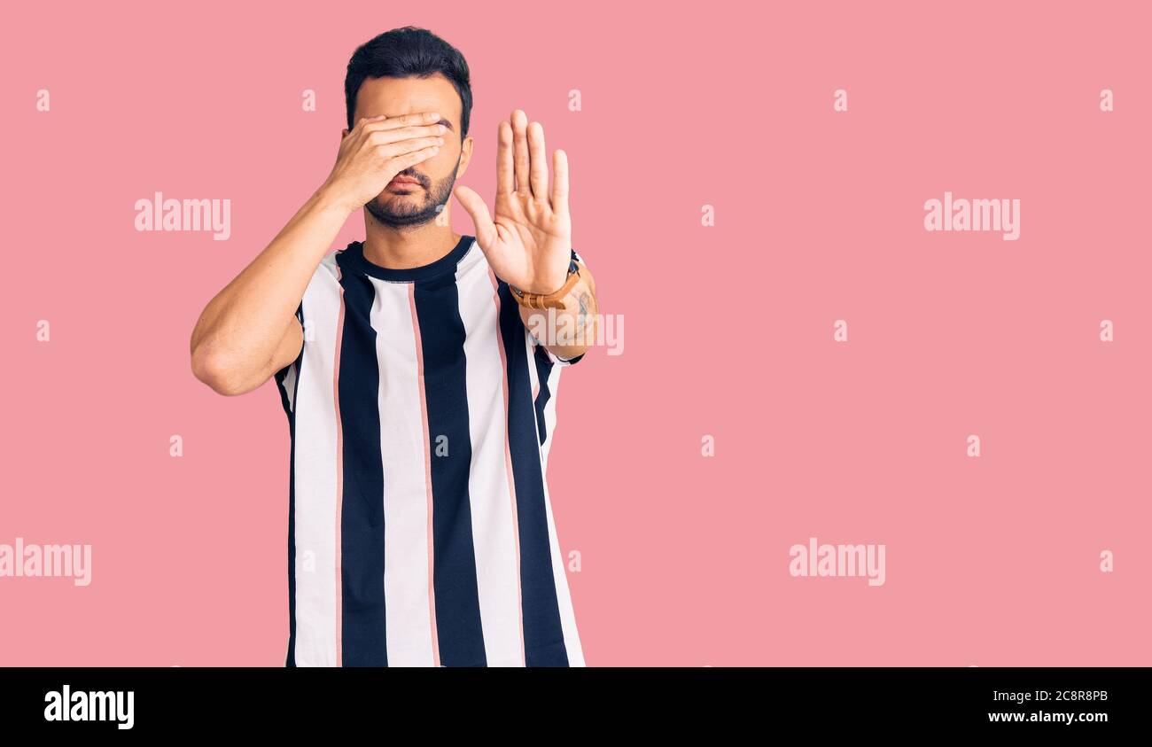 Young handsome hispanic man wearing striped tshirt covering eyes with hands and doing stop gesture with sad and fear expression. embarrassed and negat Stock Photo