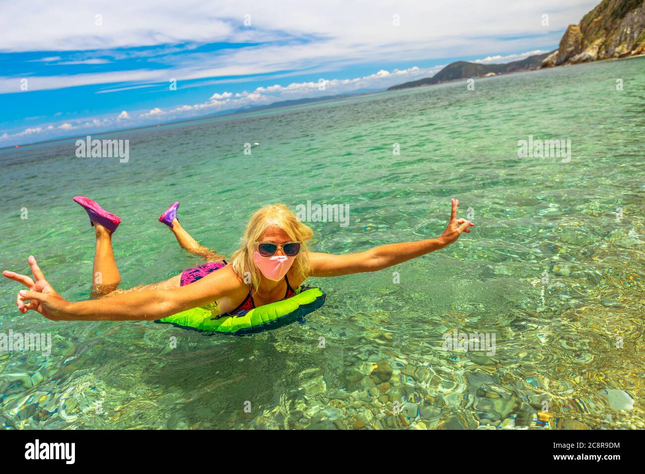 Woman swimming in Prunini beach on air mattress with surgical mask during Covid-19. Blonde woman having a bath in Elba island. Summer travel with Stock Photo