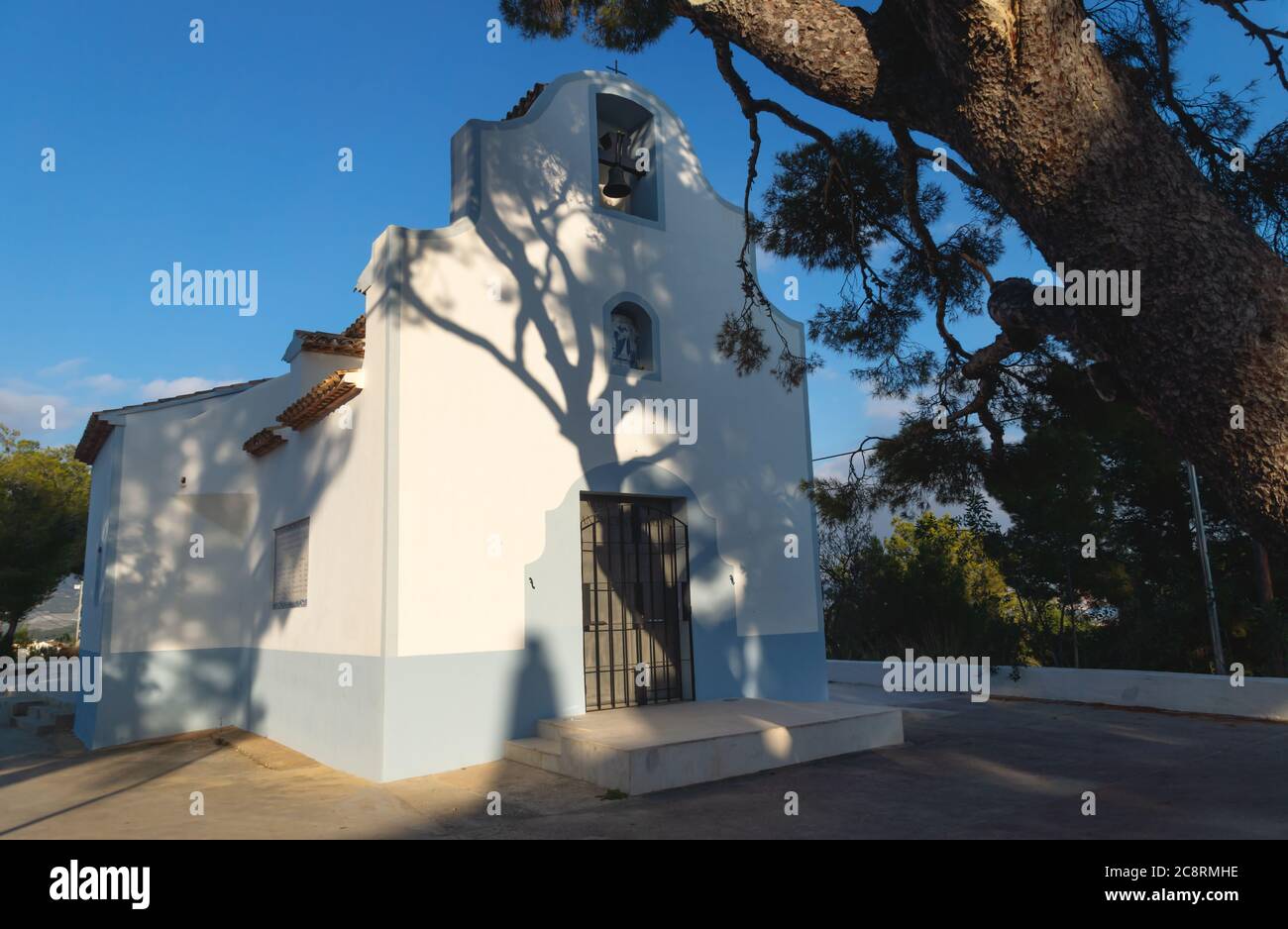 White chapel 'Ermita de Sant Vicent' sunlit with shadows from big old tree in La Nucia, Costa Blanca, Spain Stock Photo