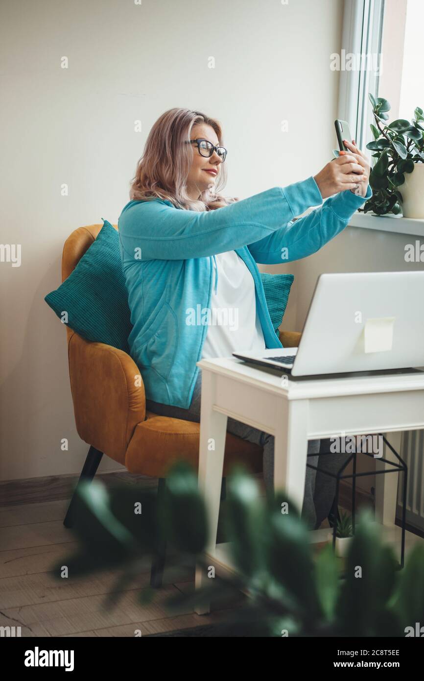 Senior caucasian businesswoman with eyeglasses making a selfie sitting in armchair and using a laptop Stock Photo