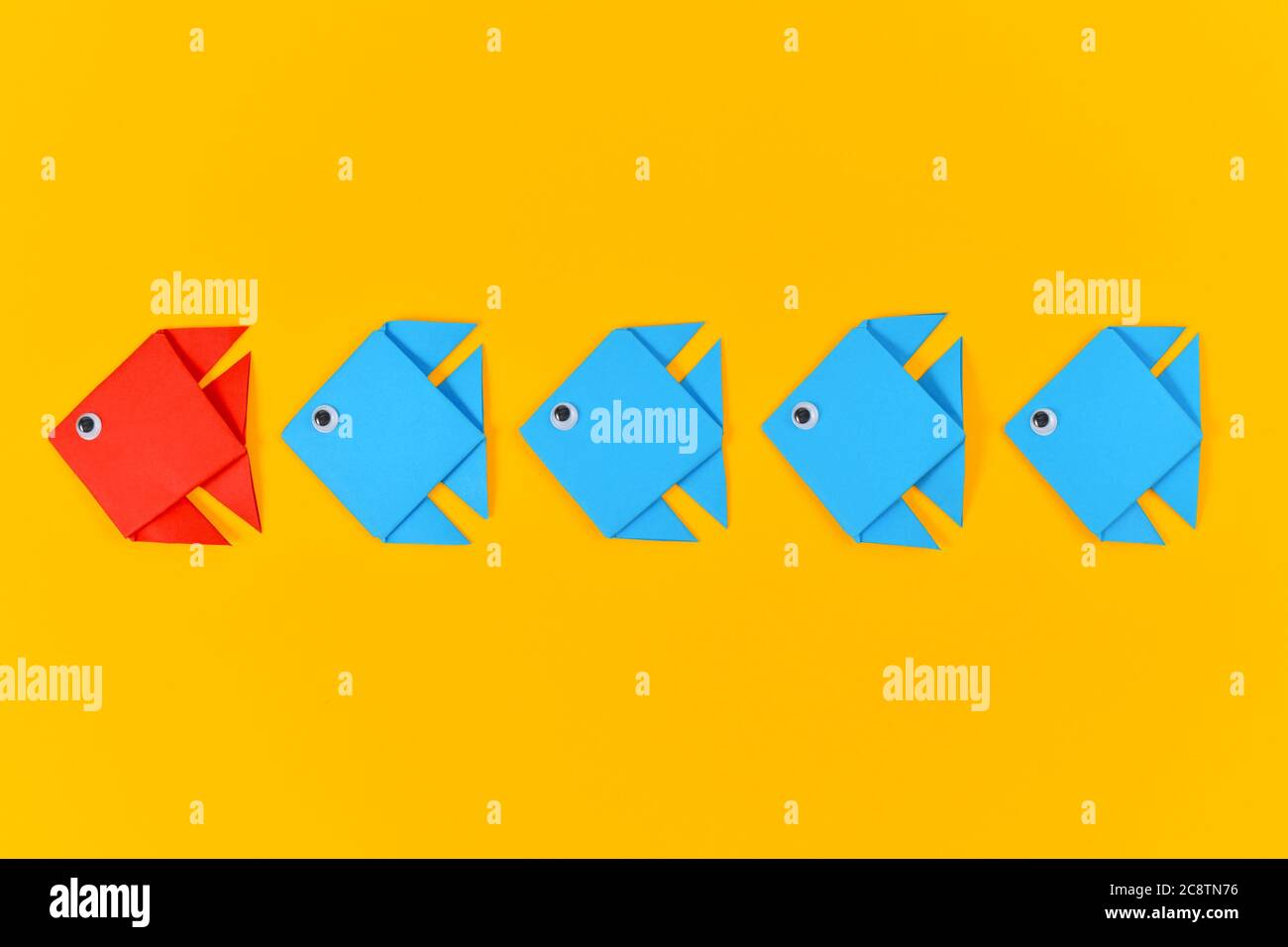 Red origami paper fish swimming in front of rows of blue fish on yellow ...