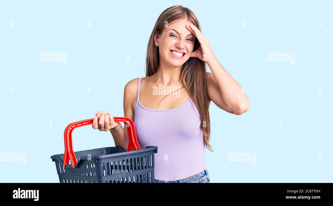 Young beautiful blonde woman holding supermarket shopping basket stressed and frustrated with hand on head, surprised and angry face Stock Photo