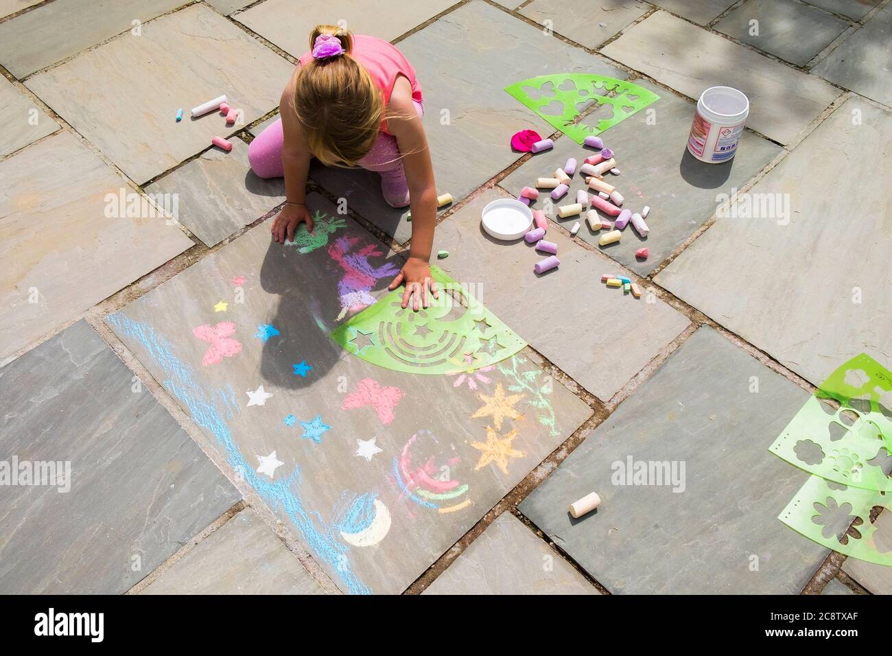 A five year old girl amusing herslef busily using plastic stencils to chalk shapes on a flagstone floor. Stock Photo