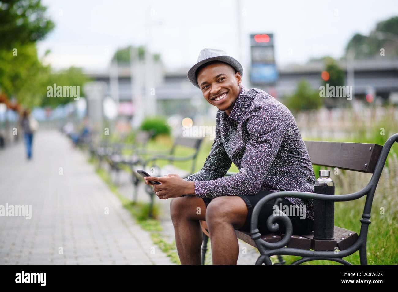 Cheerful young black man commuter sitting on bench outdoors in city ...