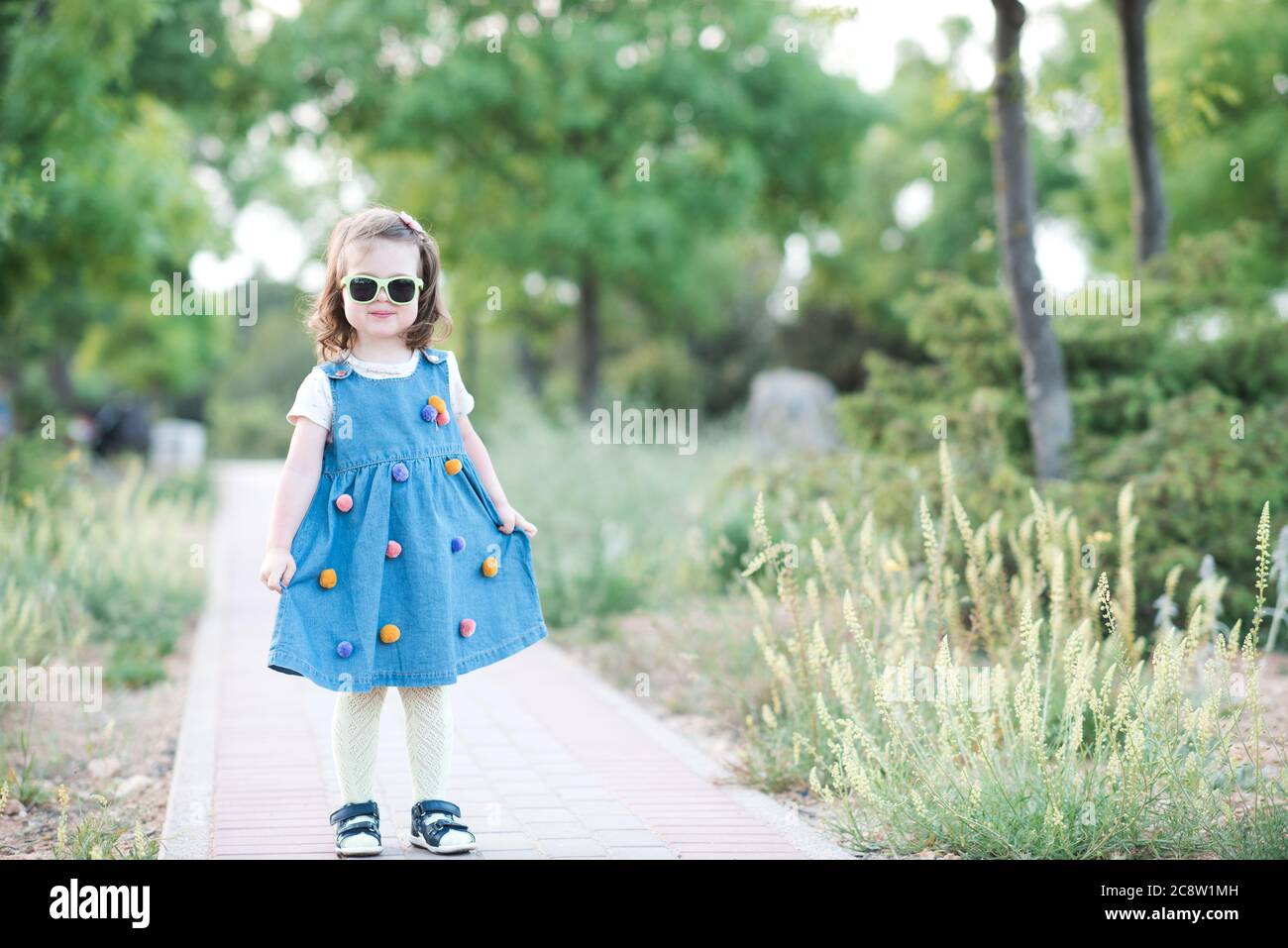 Stylish baby girl 2-3 year old wearing denim dress posing in park closeup. Looking at camera. Childhood. Summer season. Stock Photo