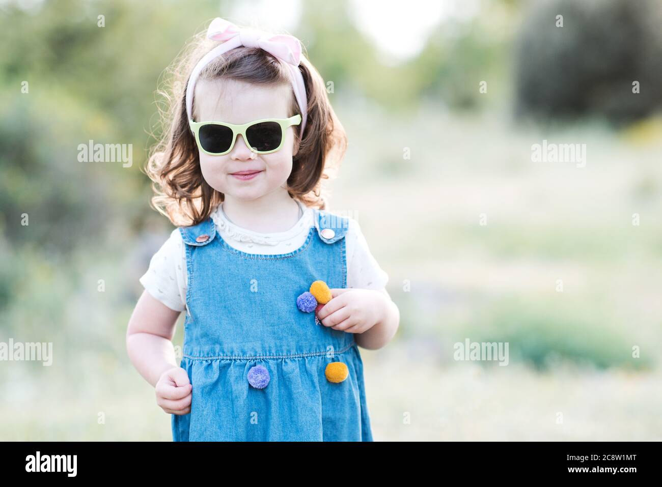 Smiling kid girl 2-3 year old wearing sun glasses and denim dress outdoors over green nature background closeup. Looking at camera. Childhood. Summer Stock Photo