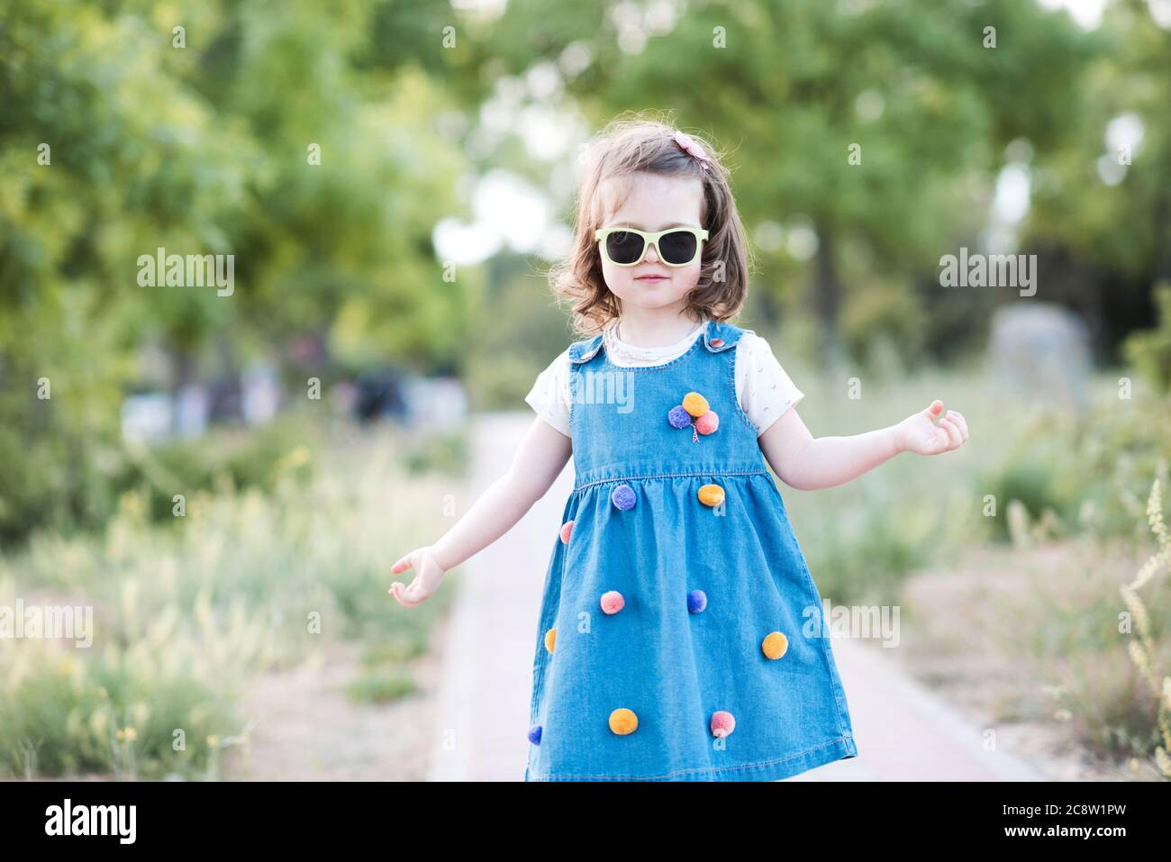 Stylish baby girl 2-3 year old wearing denim dress and sun glasses outdoors in park. Looking at camera. Summer season. Stock Photo