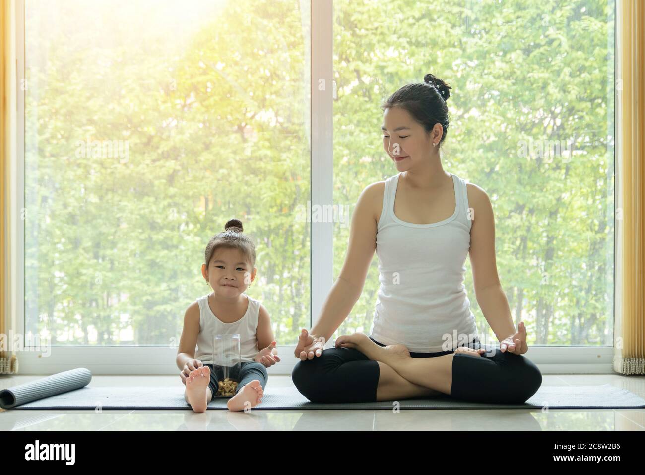 Asian mother practiced yoga by sitting in lotus position at home with a daughter sitting beside and enjoy eating snack with smiling face while workout Stock Photo