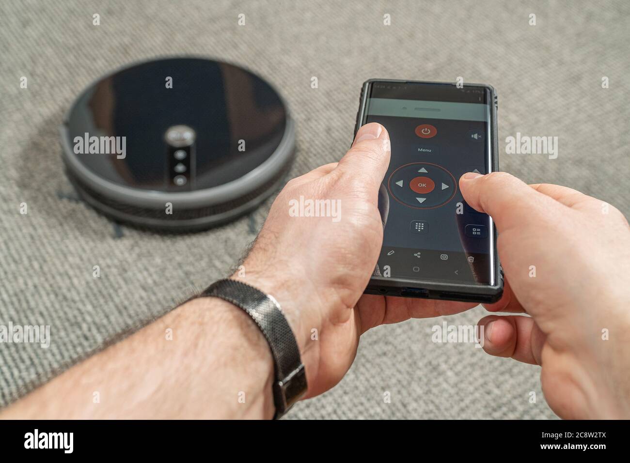 Male hands are holding the phone and through wifi controls the robot vacuum cleaner Stock Photo