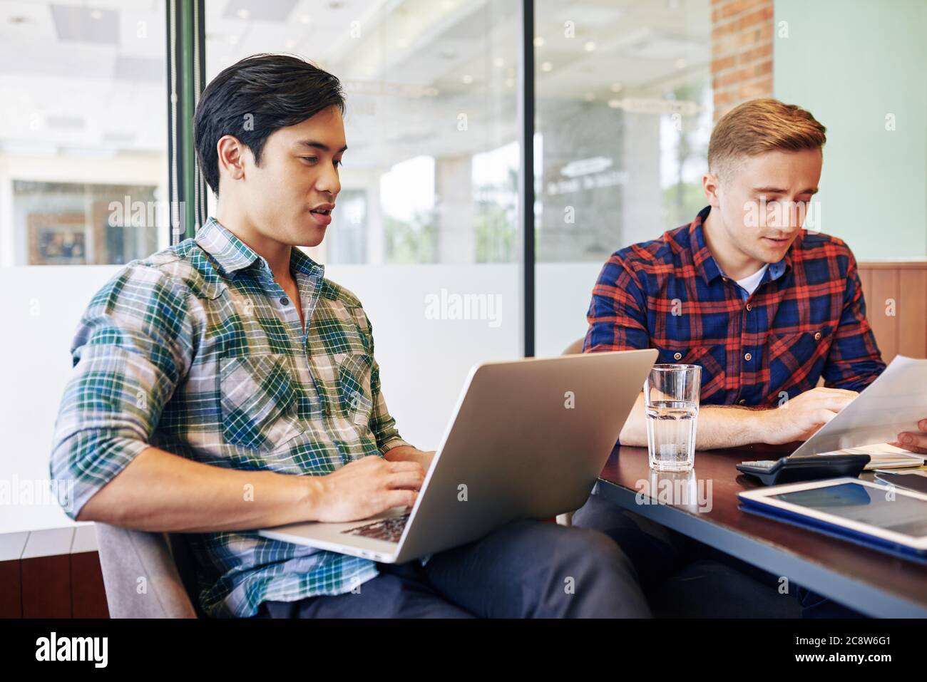 Vietnamese young man checking e-mail on laptop when his coworker reading report Stock Photo