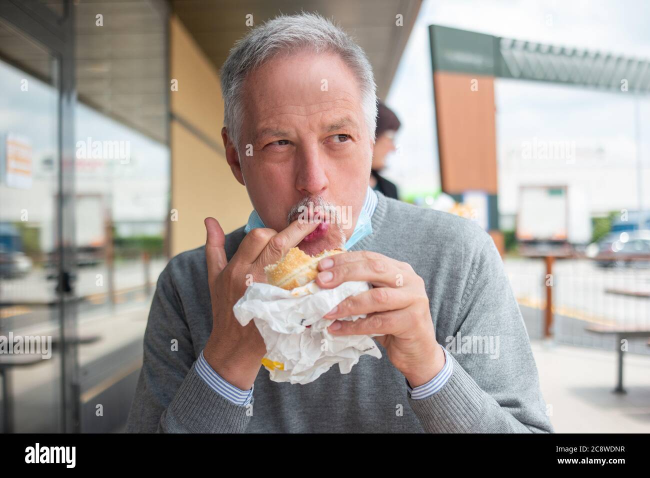 Man eating his fast food lunch hamburger during with a mask on his chin, coronavirus concept Stock Photo