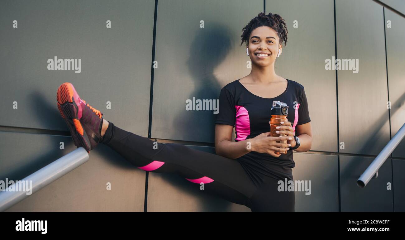 African american young woman in sportswear with wireless headphones and fitness tracker holding bottle of water and stretch leg Stock Photo