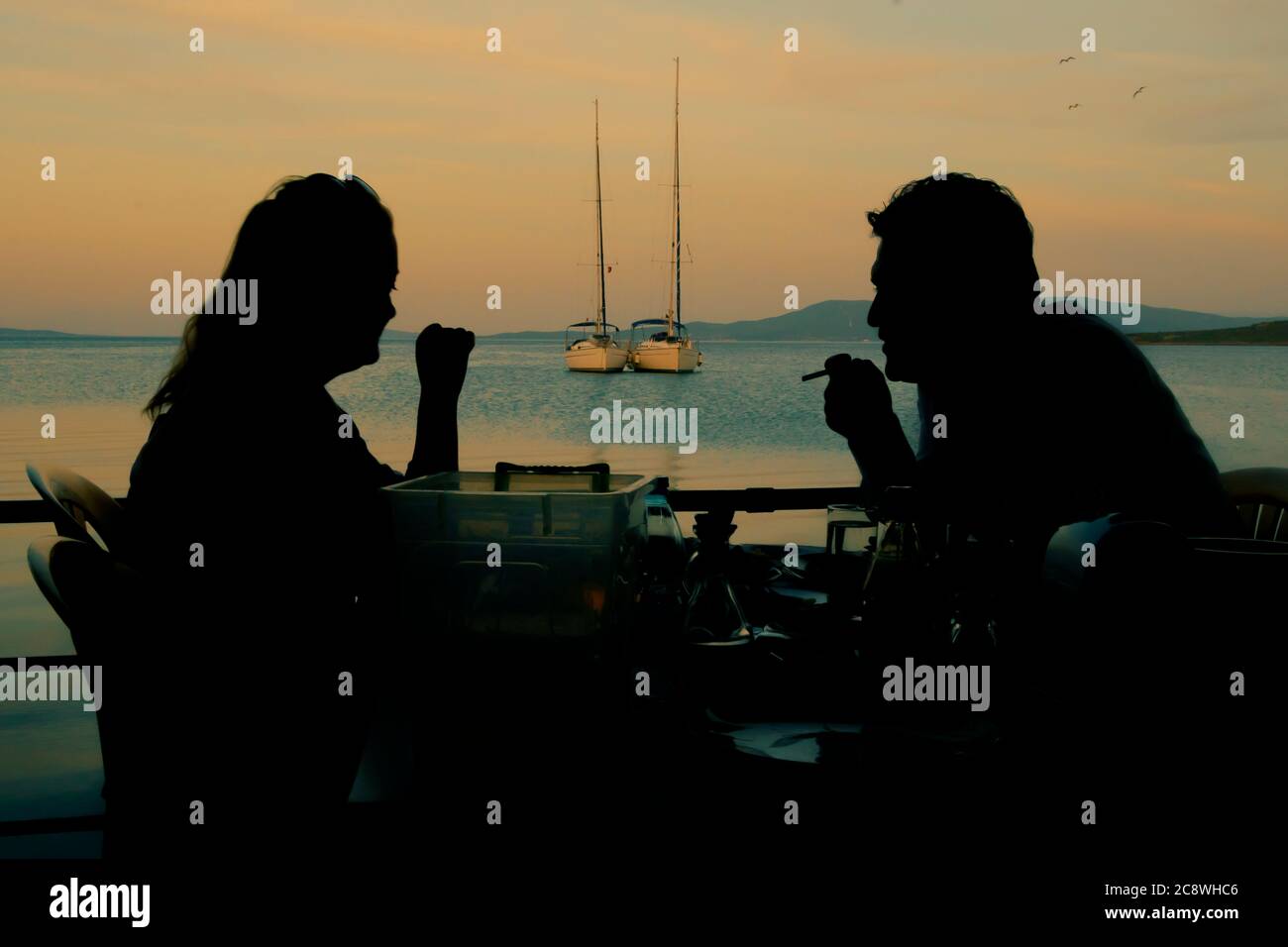 A couple having dinner in sunset time at sea side with sailboats and sea view. Stock Photo