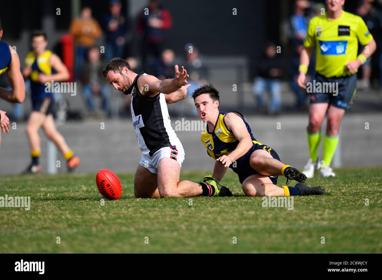 A Benalla Saints' football player stretches his arms away from the football to avoid a penalty from the onlooking umpire whilst a Mansfield opponent s Stock Photo