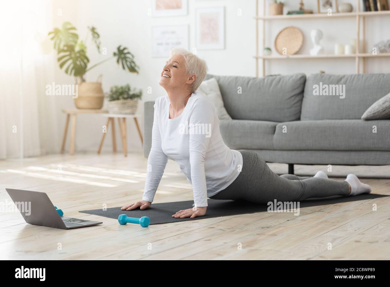 Online Yoga. Senior Woman Standing In Cobra Pose In Front Of Laptop Stock Photo